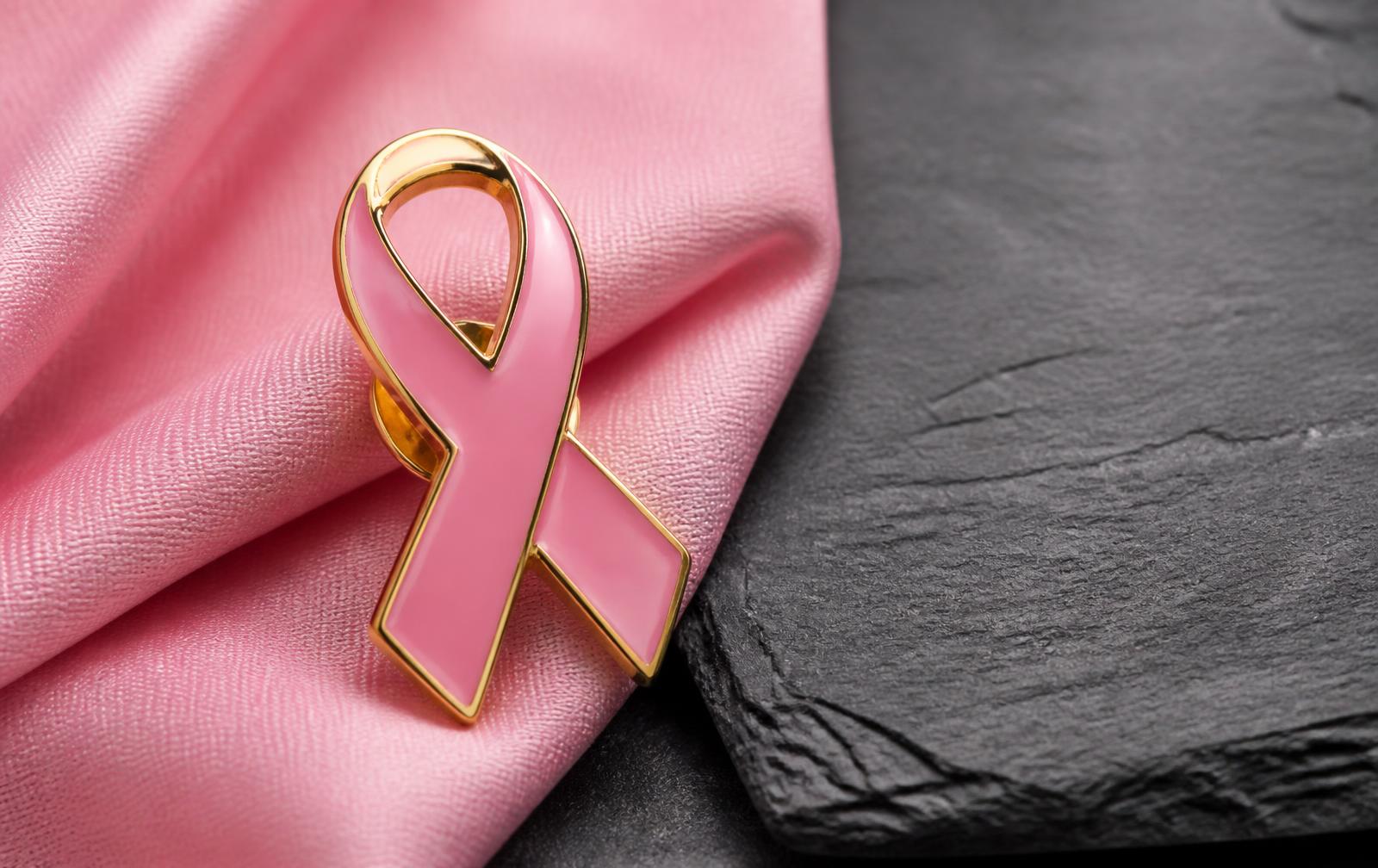 Pin metálico em formato de laço rosa com acabamento dourado sobre tecido rosa